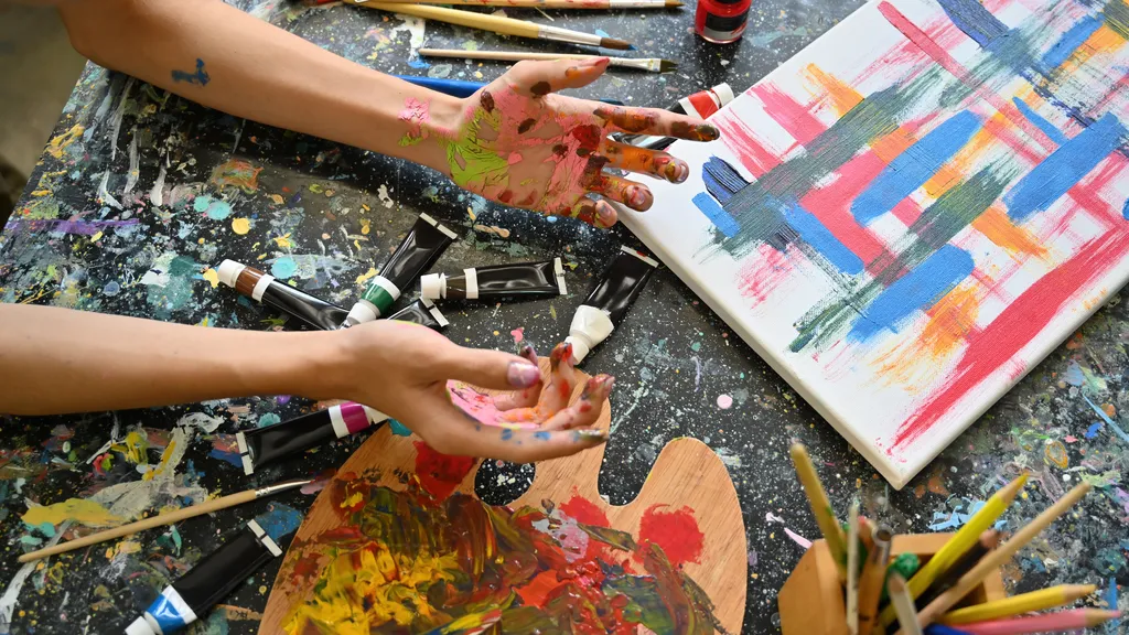 Close-up of an artist’s paint covered hands above a colourful palette and canvas